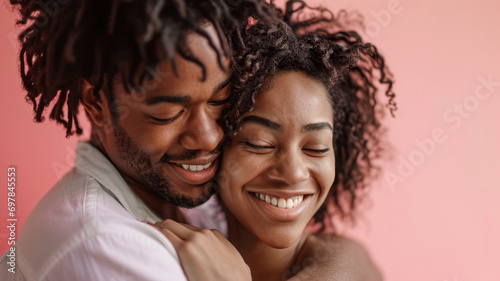  A young couple tenderly embraces against a pastel background. Their smiles radiate joy, while the light pink backdrop emphasizes their love in this intimate moment.