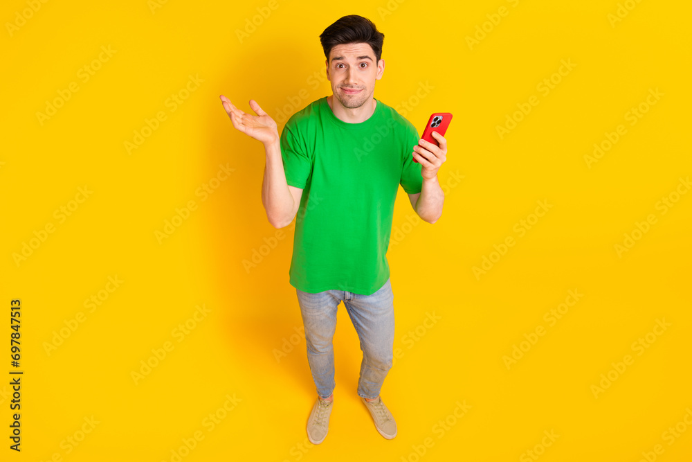 Full length photo of clueless man dressed green t-shirt jeans holding smartphone shrug shoulders isolated on yellow color background Full length photo of clueless man dressed green t-shirt jeans holding smartphone shrug shoulders isolated on yellow color background