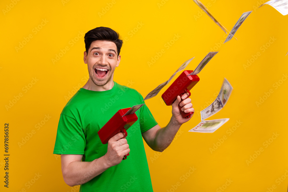 Photo of funky successful man with stylish haircut dressed green t-shirt hold guns shooting money isolated on yellow color background Photo of funky successful man with stylish haircut dressed green t-shirt hold guns shooting money isolated on yellow color background