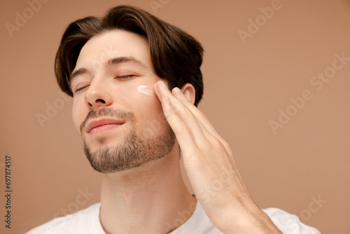 Close up of smiling man applying face cream, posing in studio. Isolated on beige background. Skin care, cosmetic, spa treatment concept