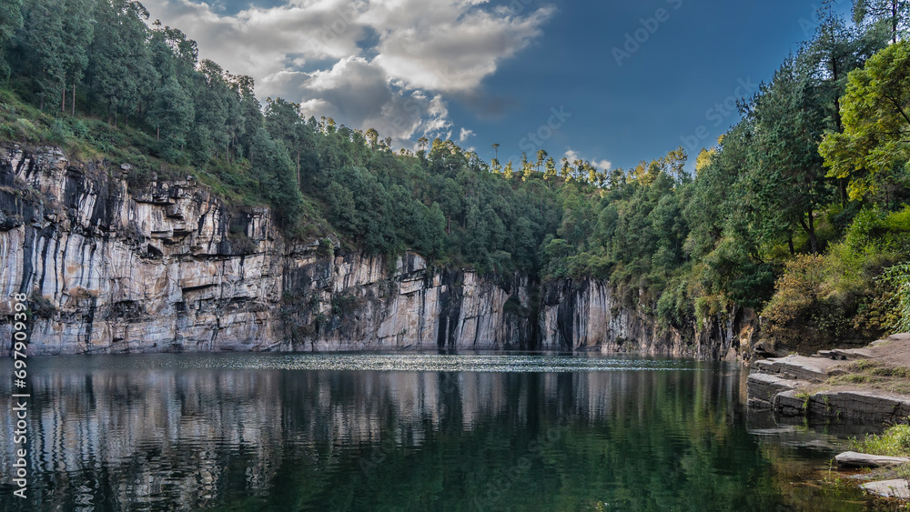 A beautiful lake in the crater of an extinct volcano. Coniferous forest grows on steep rocky shores. Blue sky with clouds . Reflection on the calm emerald water. Madagascar. Tritriva Lake.
