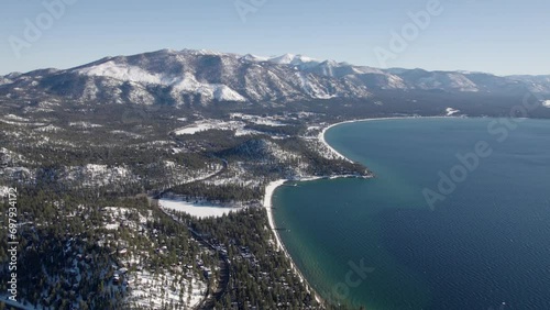 A high-flying, 4K drone shot over Lake Tahoe, California, during the winter season. The camera slowly reverses along the coastline, showing the snow-covered Sierra Nevada's in the distance.