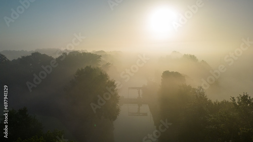 This evocative image captures the delicate balance of light and mist at dawn, with the sun rising over a tranquil river. The river itself is barely visible, as a thick blanket of fog softly engulfs