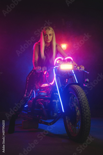Young beautiful girl in the neon lights stands near the motorbike.