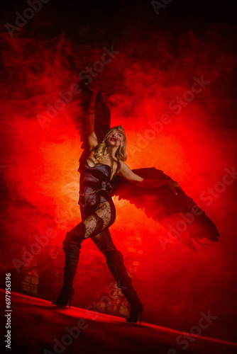 Beautiful girl an angel with the black wings posing in the smoke on dark background in the red light. Angel of the night concept.