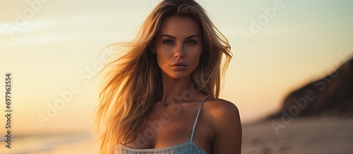 Blonde woman in alluring swimwear posing on beach at sunset. Perfect wavy hair, tan body. Tropical summer vibe. photo