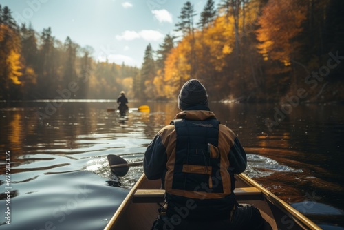 Person canoeing on a tranquil river with autumn foliage and a fisherman in the background. © robertuzhbt89