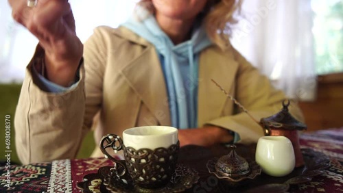 Woman pouring Turkish coffee from cezve into cup. Closeup slow motion shot of female hand with cup on square plate, on table in cafe outdoor. Traditional hot unfiltered coffee served in restaurant photo