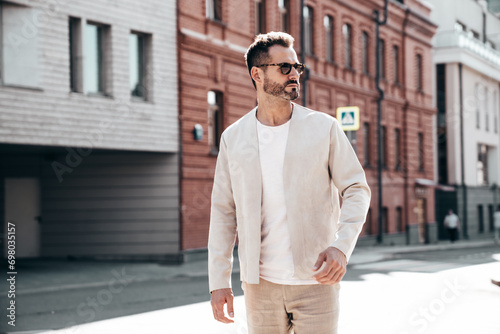 Handsome confident stylish hipster lambersexual model. Sexy modern man dressed in elegant white suit jacket. Fashion male posing in the street background in Europe city at sunset. In sunglasses