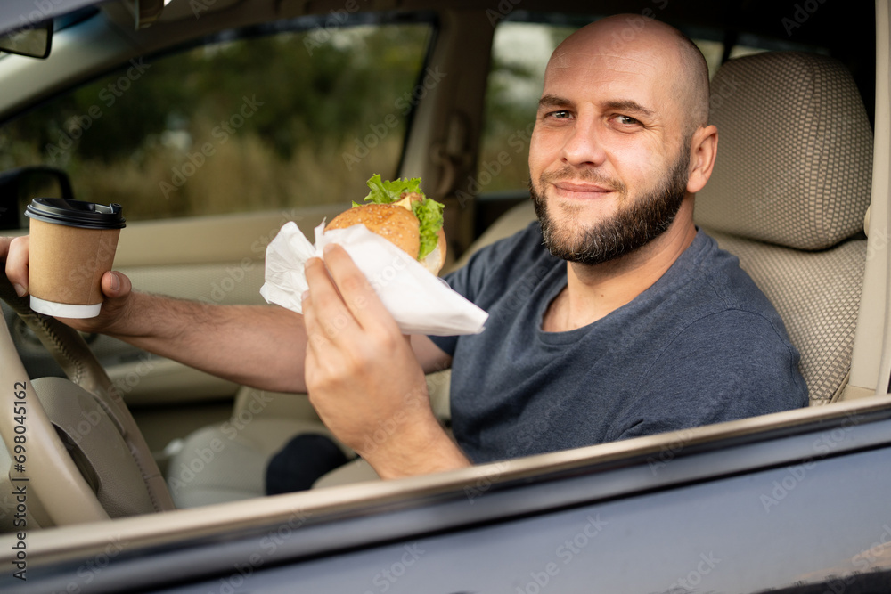 A bald smiling man driving a car drinking takeaway coffee and eating a burger looks out the window. Dangerous driving - obrazy, fototapety, plakaty 