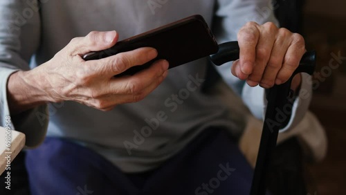 impersonal elderly man sitting with a walking cane watching video hosting using a smartphone holding it horizontally. daily life of old people photo