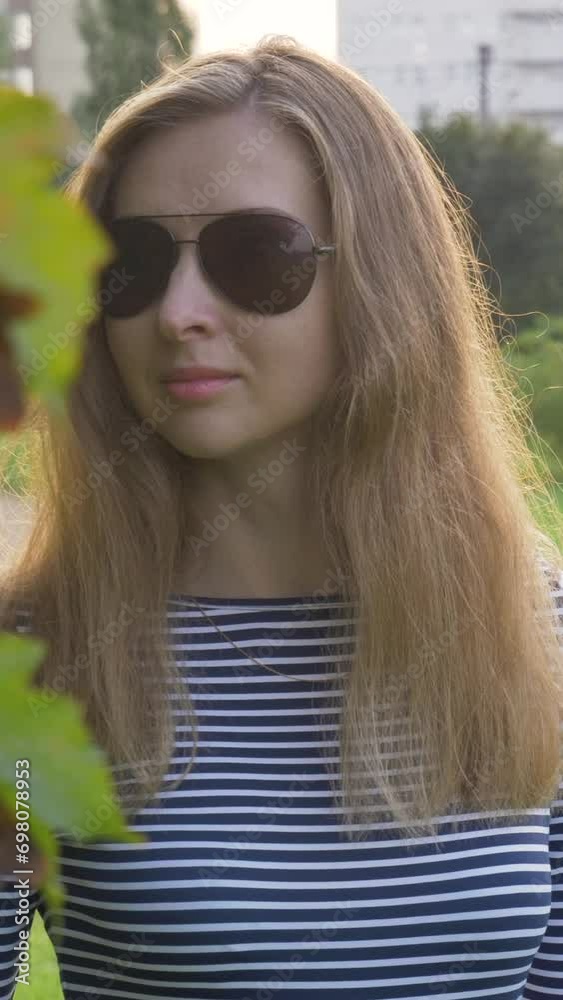 Young caucasian woman with long hairs in sunglasses is walking near tree branch with leaves in park. Vertical Video Young caucasian woman with long hairs in sunglasses is walking near tree branch with leaves in park. Vertical Video