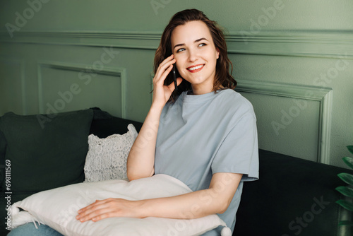 A smiling pretty young woman makes a call on her smartphone while enjoying a conversation in a gray living room. Joyful Chat. Pretty Woman Smiling during Smartphone Call.