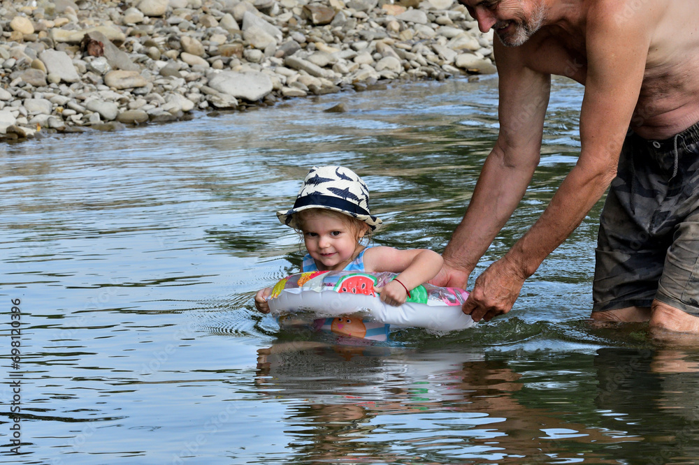A grandfather teaches his granddaughter to swim in the river using an inflatable lifeline