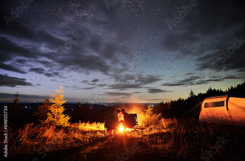Romantic couple enjoying at starry sky. Young people sitting in chairs and warming by campfire near tourist tent. Travelling couple having a rest together. Night camping.
