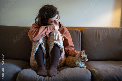 Upset sad depressed teenager sitting on sofa, putting head on knees, stroking beloved breed cat Devon Rex. Teen girl feeling lonely, stressed in need of friendship, emotional support, tactile contact