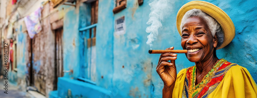 Elderly Woman Enjoying a Cigar Against a Blue Wall in Havana. Smiling Senior Lady in Yellow Dress and Straw Hat with Cigar in Cuba photo