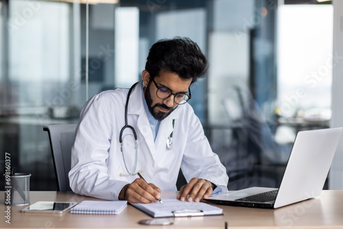 Concentrated young Indian male doctor in white coat sitting in office at table with laptop and writing medical results and documents photo
