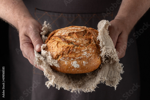 Man baker holding fresh spelt wheat loaf of bread on rustic linen towel on dark background. photo