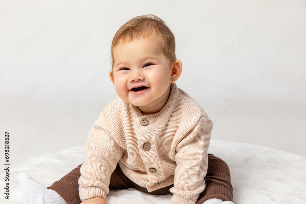 Happy smiling adorable newborn baby girl in beige and brown suit sitting on light white home carpet on white background. 6 to 7 months old development. advertising of toys and goods for children Happy smiling adorable newborn baby girl in beige and brown suit sitting on light white home carpet on white background. 6 to 7 months old development. advertising of toys and goods for children