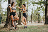 Fit girls in a city park after training, holding a ball and rackets, having a conversation. They enjoy a break in the natural environment, motivated to maintain a healthy lifestyle.