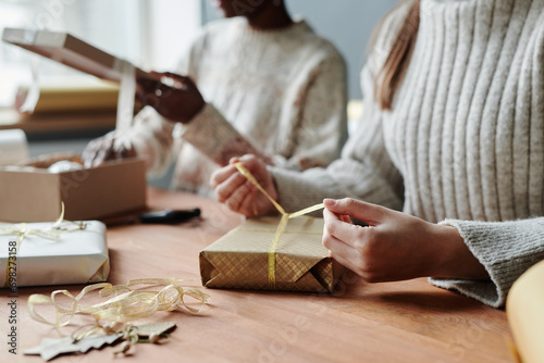 Hands of young unrecognizable woman in sweater tying ribbon around giftbox wrapped into craft paper while preparing presents with friend