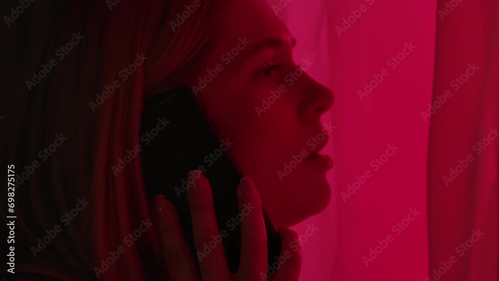 A concerned woman standing near a window illuminated by red and blue police lights. Woman talking emotionally on a smartphone, close up. A concerned woman standing near a window illuminated by red and blue police lights. Woman talking emotionally on a smartphone, close up.