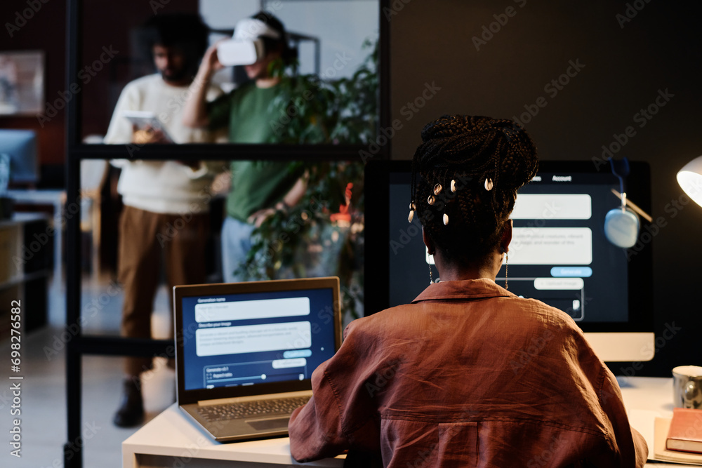 Rear view of young African American female designer sitting in front of laptop and desktop computer screens with AI design tasks Rear view of young African American female designer sitting in front of laptop and desktop computer screens with AI design tasks