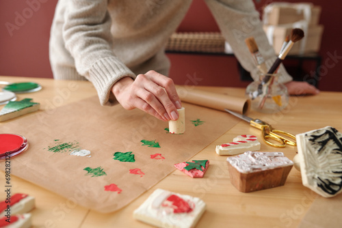 Hand of young unrecognizable woman making prints of Christmas symbols on paper while bending over table with various supplies