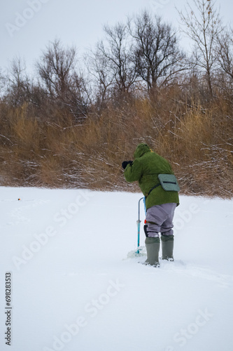 A fisherman is fishing in an ice hole on a frozen pond. The joy of winter fishing. A fisherman on a winter fishing trip on a cloudy day. 