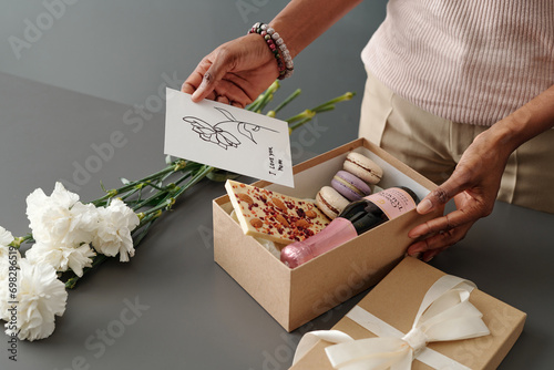 Woman putting handmade postcard with greetings for Mother Day into giftbox with white chocolate, macaroons and bottle of champagne