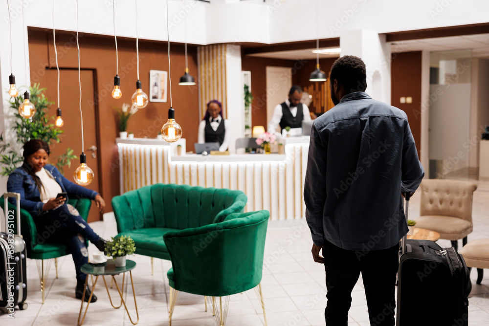 Fototapeta premium African American man tourist arriving at hotel, standing with luggage in foyer or lobby, rear view. People tourist arrive at resort waiting for check-in procedure, holiday season