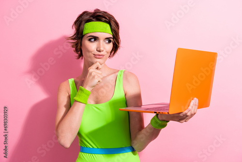 Photo portrait of pretty young girl touch chin hold orange netbook dressed stylish green sport jumpsuit isolated on pink color background