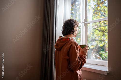 Lonely teenager standing near window, looking at sunny summer weather with sadness, sorrow, spending time alone at home. Unsociable teen girl having no friends. Youthful crisis, communication problems photo