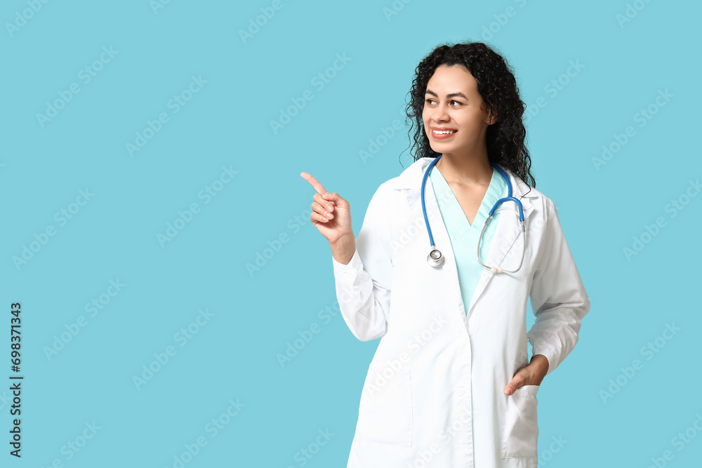 Beautiful young happy African-American female doctor with stethoscope pointing at something on blue background Beautiful young happy African-American female doctor with stethoscope pointing at something on blue background