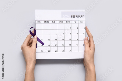 Female hands with purple ribbon and paper calendar on grey background. Cancer awareness concept