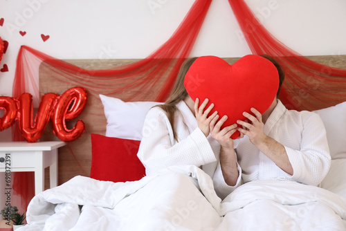 Young couple hiding behind heart-shaped pillow in bedroom on Valentine's Day