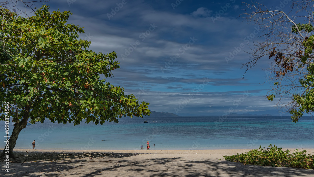 A beach on a tropical island. Tiny figures of people at the water's edge. Boats in the turquoise ocean. Shadows of trees on the sand. Blue sky with clouds. Madagascar. Nosy Tanikeli 