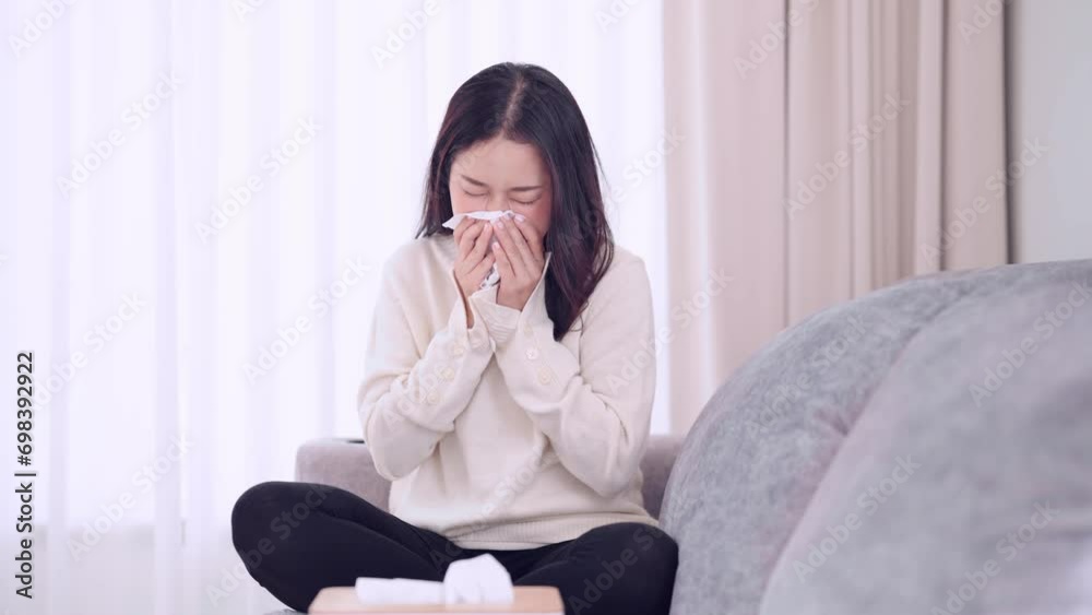 Asian woman coughing and sneezing, feeling anxious about getting sick or unwell while sitting on her home sofa. Capturing the moment of discomfort and concern at home Asian woman coughing and sneezing, feeling anxious about getting sick or unwell while sitting on her home sofa. Capturing the moment of discomfort and concern at home