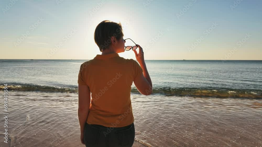 A person holding sunglasses enjoys the serene view of a sunset over a calm sea, symbolizing peace and relaxation A person holding sunglasses enjoys the serene view of a sunset over a calm sea, symbolizing peace and relaxation