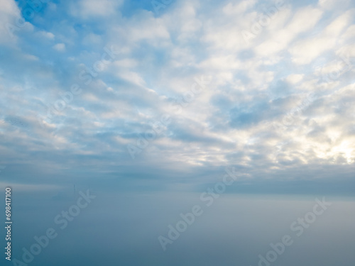 This image  captured by a drone above the mist  depicts a serene skyscape where the soft white of the altocumulus clouds meets the gentle gradient of dawn light. The blanket of mist creates a