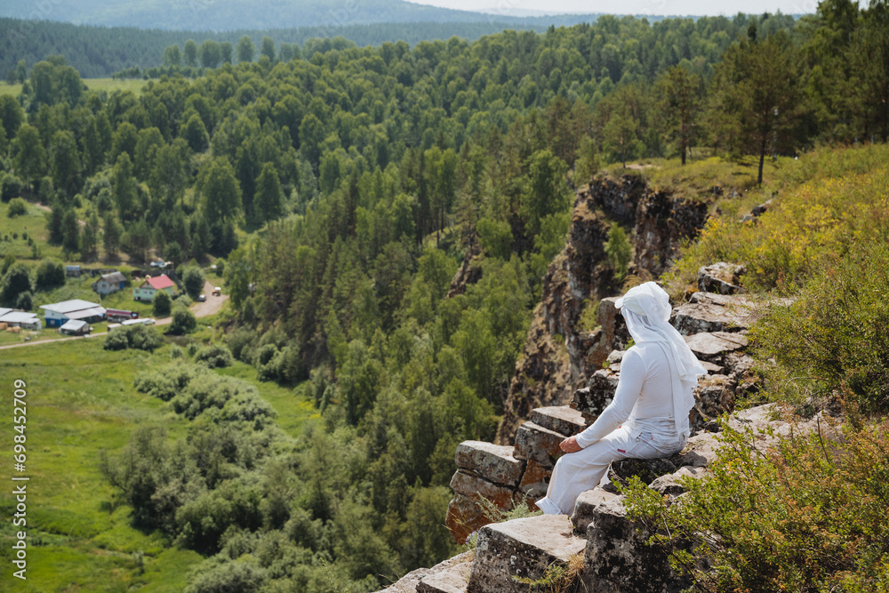 A guy in white clothes sits on the edge of a cliff, a monk meditates in nature in the mountains, a pilgrim meditates sitting on a rock high in the mountains.