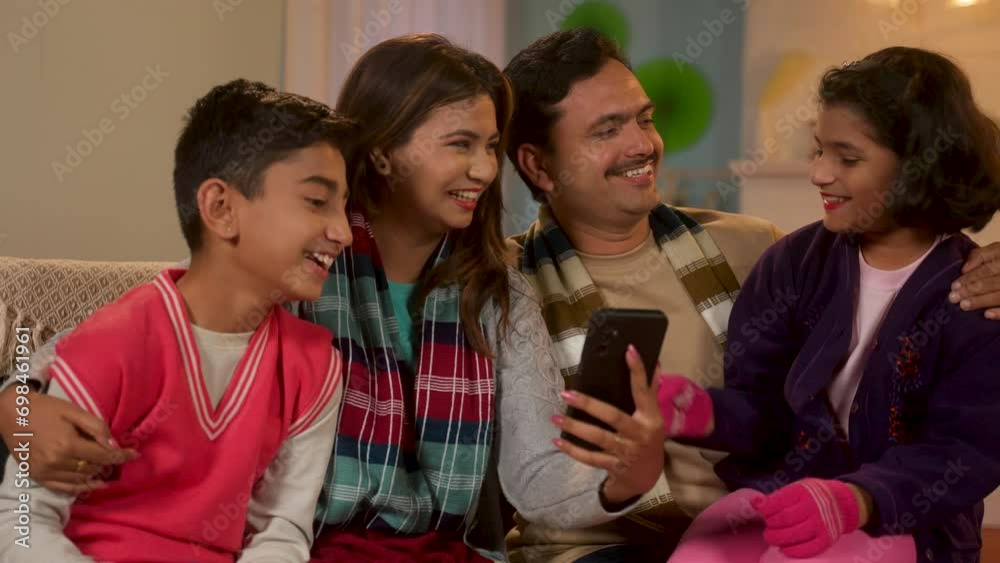 Joyful parents with kids using mobile phone with winter wear at home - concept of social media sharing, technology and weekend holidays Joyful parents with kids using mobile phone with winter wear at home - concept of social media sharing, technology and weekend holidays