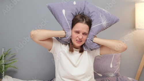 Angry sad unhappy Caucasian woman closing her ears with pillow in morning because of neighbor noise sleepless girl annoyed by alarm clock or loud neighbors photo