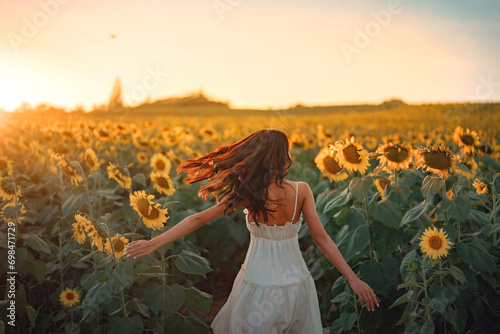 Carefree Happy beautiful young woman in white dress opened arms up in air and looking at sunset in a large field of sunflowers, Freedom concept, Enjoyment, Summer time. © oatawa