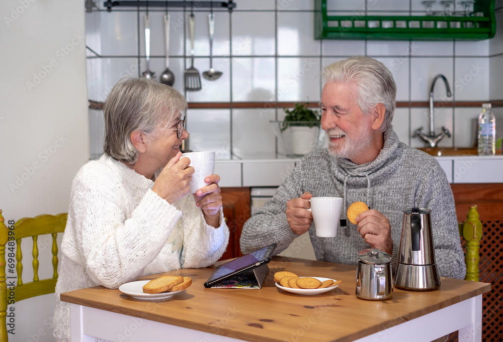 Portrait of attractive smiling senior couple having breakfast together sitting at kitchen table. Elderly 70s husband and wife enjoying relaxed retirement