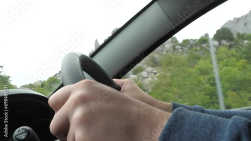 Man drives car holding steering wheel past cliffy mountains with pine wood closeup. Man enjoys car driving at highland on gloomy day. Driver controls car on road trip across picturesque mountain site photo