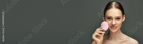 young woman holding cleansing brush on grey background, beauty gadget and skin care banner photo