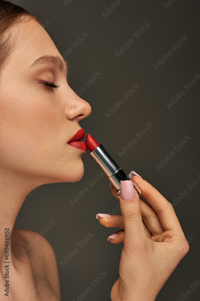 Fototapeta premium side view of beautiful young woman applying red lipstick on grey background, beauty and makeup