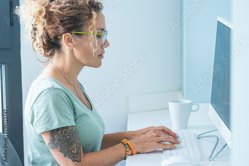 Cute modern style adult woman tattooed write and work on desktop computer in office or home room - concept of free people and online technology job activity - pretty adult lady use keyboard in white